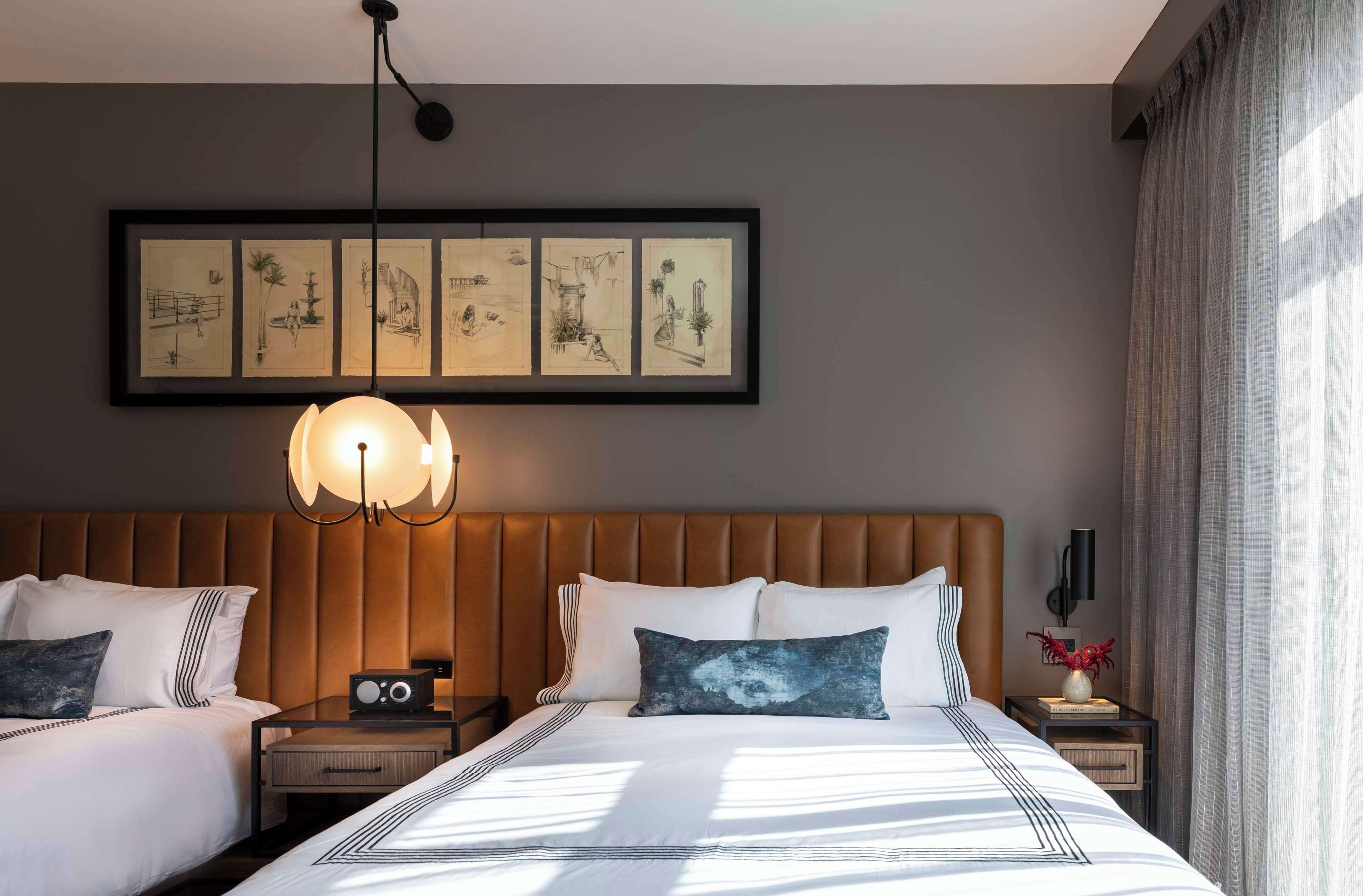 A hotel room with two beds, a large framed artwork above the beds, a modern light fixture, and a nightstand with a radio and decorative items.