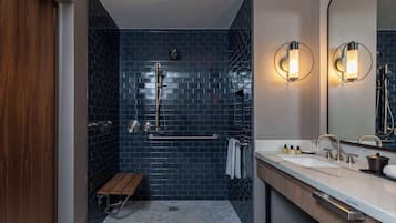 Une salle de bain moderne avec une douche carrelée de carreaux bleu foncé, un banc en bois, un lavabo blanc avec des robinets dorés et deux appliques murales.