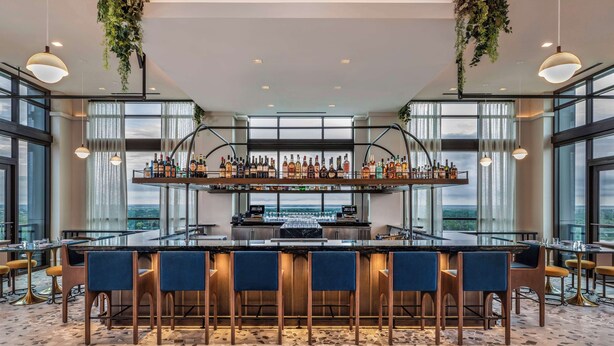 Una zona de bar con una barra larga, varias estanterías llenas de botellas y taburetes.