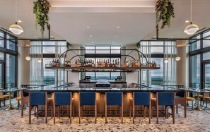 Una zona de bar con una barra larga, varias estanterías llenas de botellas y taburetes.