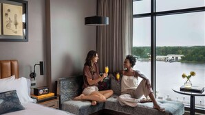 Habitación de hotel moderna con un ventanal grande, un sofá, una mesita de noche con una radio y vistas a un río.
