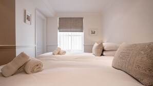A hotel room with a large bed, two pillows, a window with blinds, and a door. - 2 South Beach Court - Beach Apartment (Tenby)