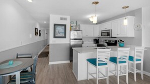 A modern kitchen with white cabinets, a stainless steel refrigerator, a microwave, and a stove. There is a dining area with a table and chairs. - Gulf Front in Seacrest, Beautiful Views & Easy Beach Access + Free Tickets! (Seacrest)