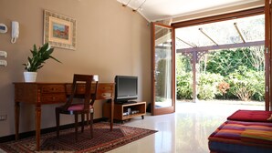 Una sala de estar con un escritorio de madera, un televisor, una silla, un sofá rojo, una planta en maceta y un amplio ventanal que da a un jardín.