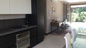 Una cocina moderna con armarios negros, encimera de granito y electrodomésticos de acero inoxidable. Hay un comedor con una mesa y sillas de vidrio, y vistas a un jardín a través de una puerta de cristal.