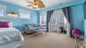A bedroom with a bed, a gray sofa, a wooden bench, a large window, and a balcony. - Ocean view, Walkable to Everything in the heart of Carolina Beach (Carolina Beach)
