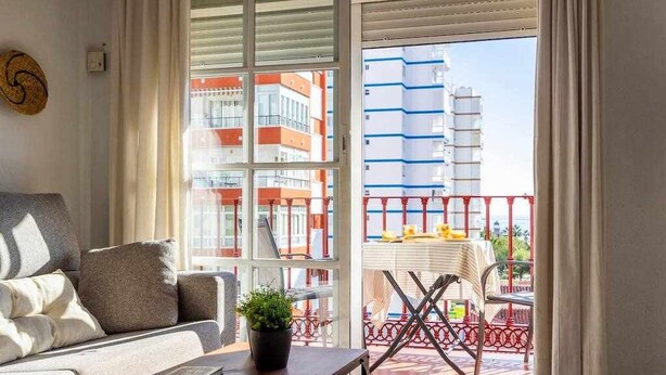 En stue med en sofa, et sofabord med en bog og en plante, samt udsigt til en balkon med et bord og stole.