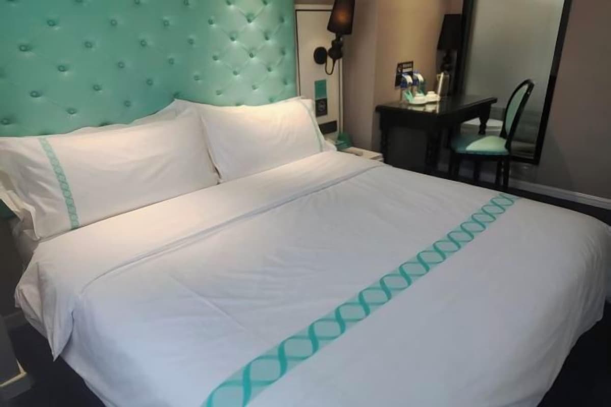 Una cama bien tendida con ropa de cama blanca y detalles azul verdoso, un cabecero capitonado y una mesita de noche con una lámpara y un espejo pequeño.