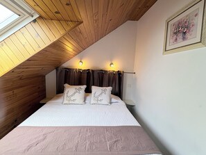 Une chambre avec un plafond en pente, des poutres en bois, un lit agrémenté d’oreillers décorés d’un motif d’ancre, deux lampes fixées au mur et un tableau encadré accroché au mur.