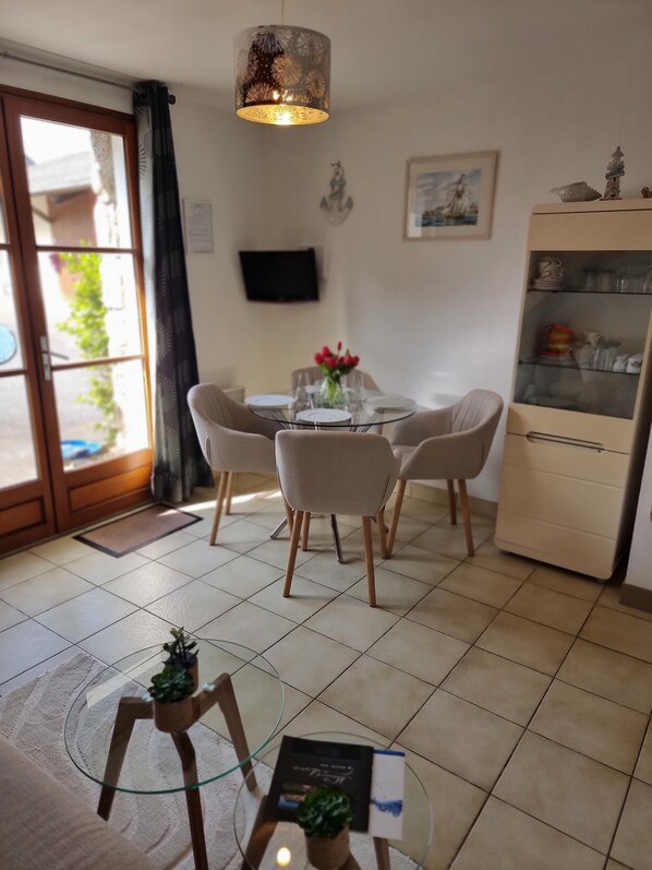 A dining area with a glass table, chairs, a decorative hanging light, a television on the wall, and a cabinet with glass doors.