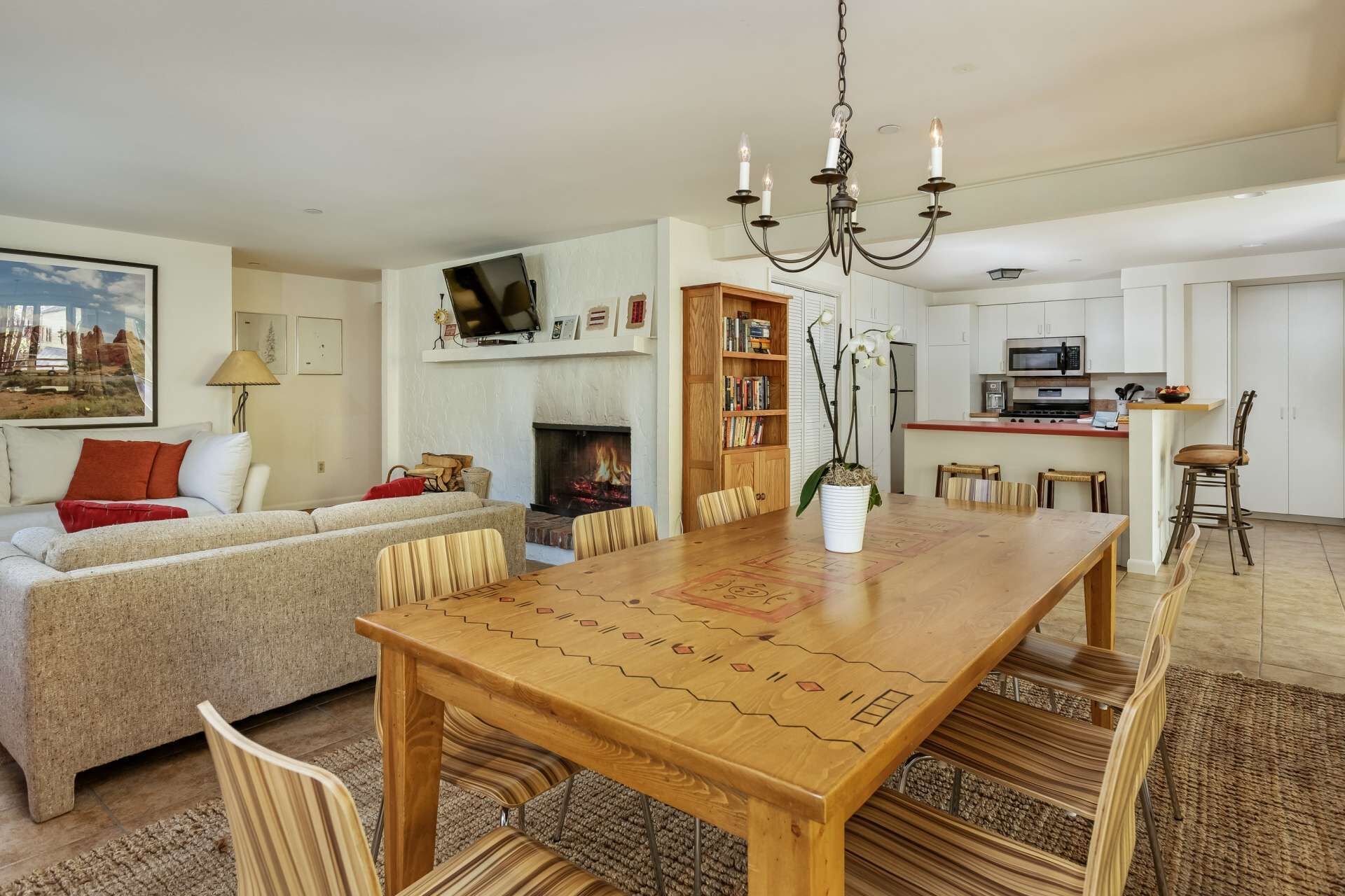 A living room with a sofa, a wooden dining table, and chairs. There is a fireplace, a bookshelf, and a kitchen area with a microwave.