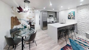 Een moderne keuken met een centraal kookeiland, een eetgedeelte met een glazen tafel en een woonruimte met een bank en een vloerkleed met dessin.