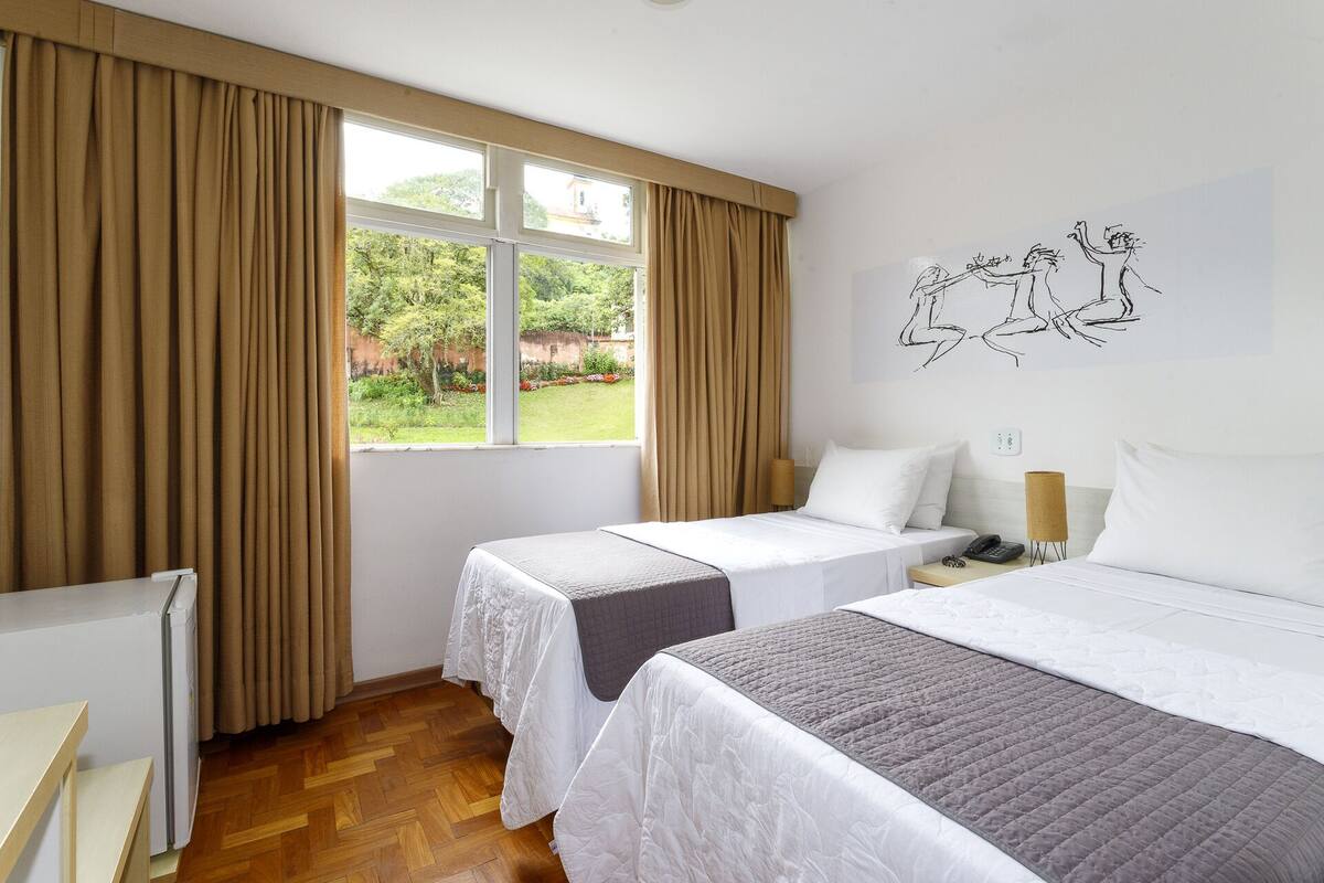 A hotel room with two beds, a large window with curtains, a wall art piece, and a bedside lamp.