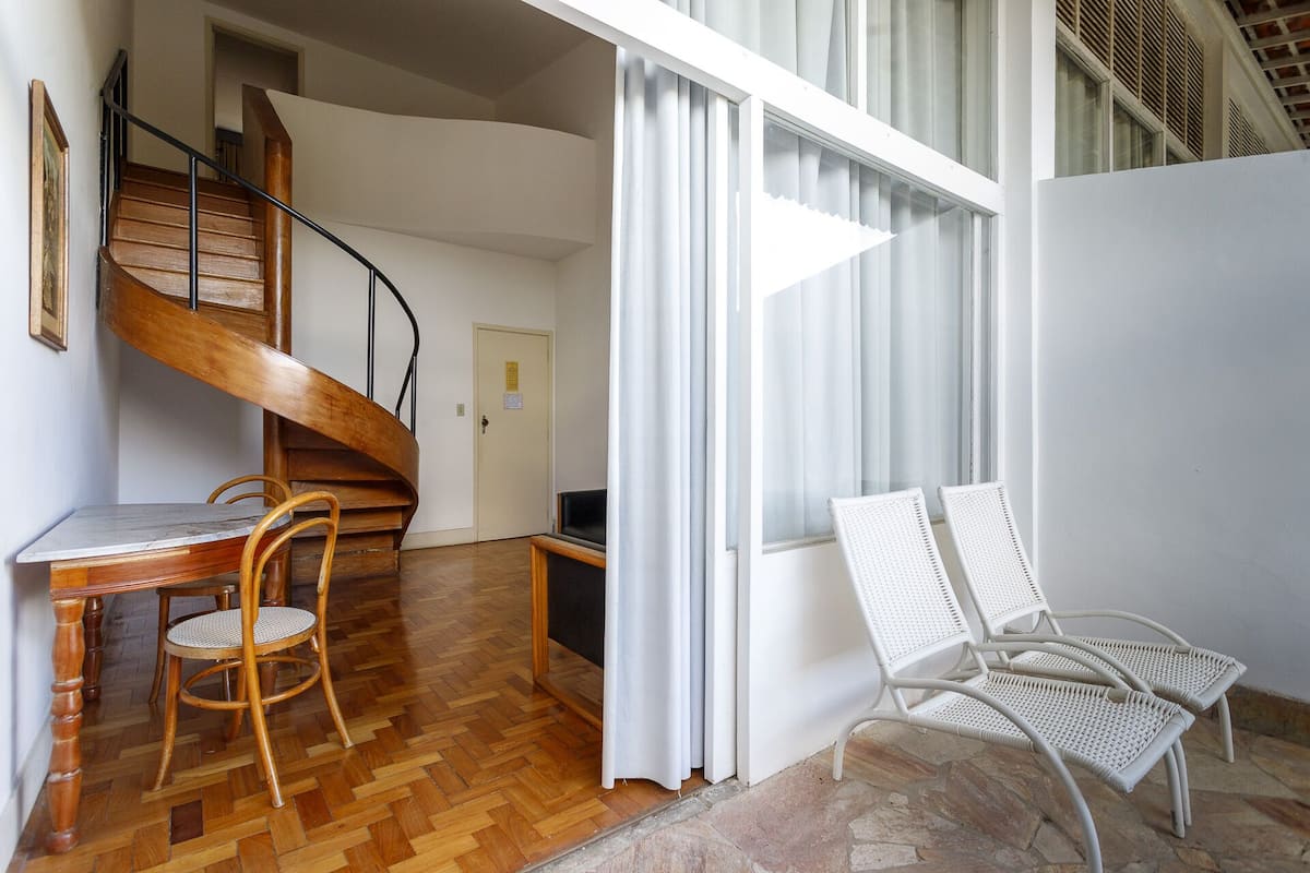 A room with a wooden staircase, a dining table with chairs, and a window with white curtains.