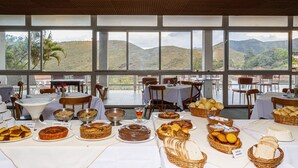 A dining area with tables set for breakfast, featuring a variety of pastries and bread, with a scenic view of hills in the background. - Grande Hotel de Ouro Preto (Ouro Preto)