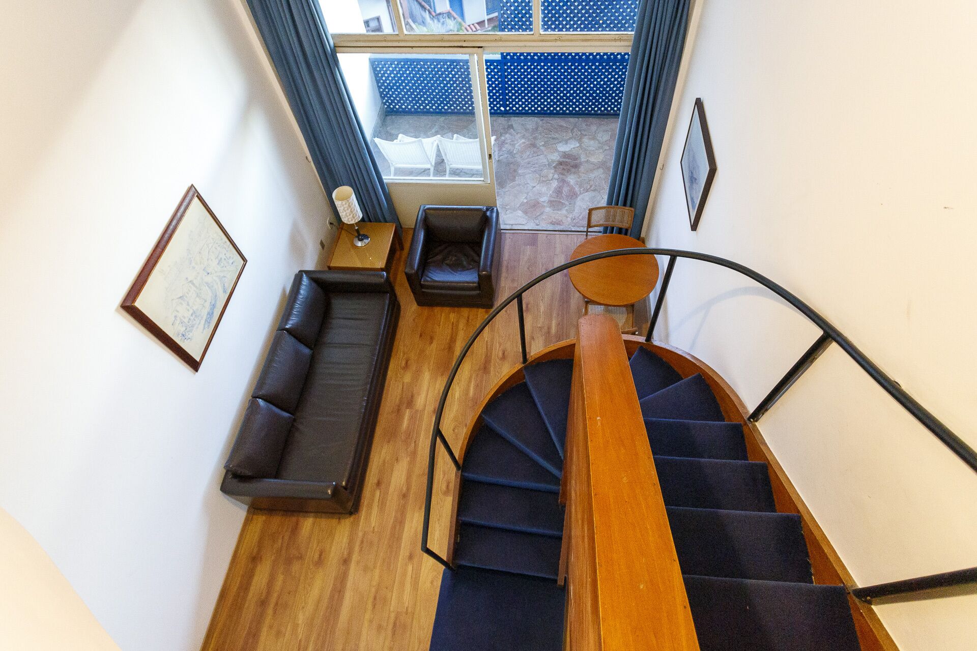 Un escalier doté d’une main courante en bois et d’une rampe en métal noir, menant à une pièce où se trouvent un canapé bleu foncé, une petite table avec une lampe et un tableau encadré accroché au mur.