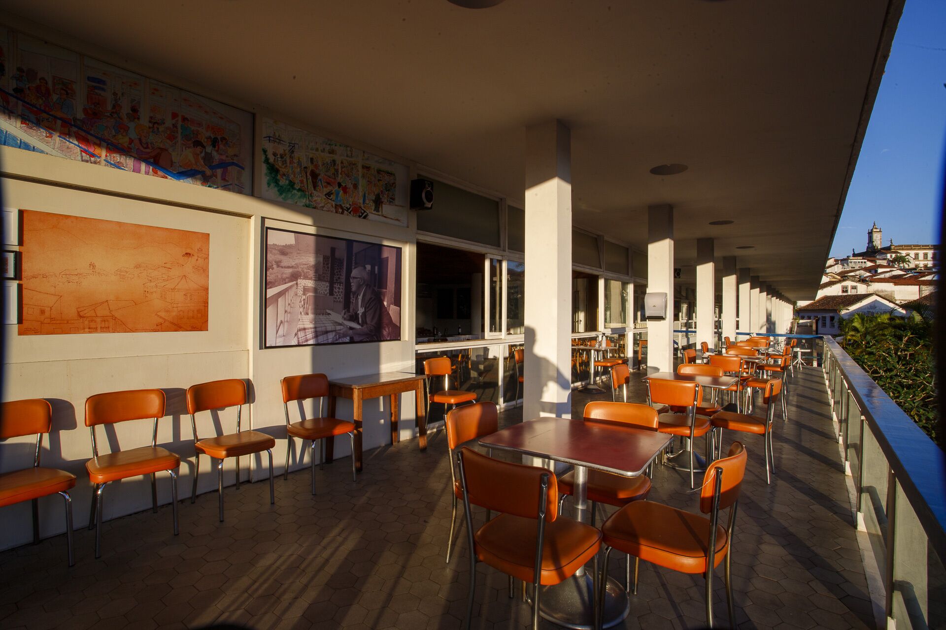 A covered outdoor dining area with orange chairs and tables, large windows, and artwork on the walls.