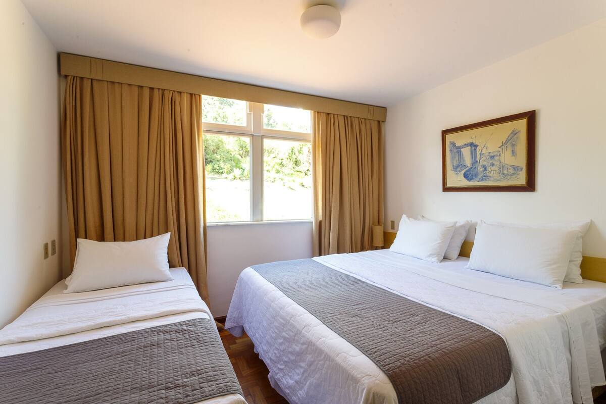 A hotel room with two beds, beige curtains, a window with a view of greenery, and a painting on the wall.