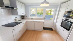 A kitchen with white cabinets, a black stove, a sink, and a window with a view of greenery. - Oceanside | Sleeps 6 |Walk to Beach & Stylish (Croyde)