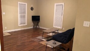 A living room with wooden flooring, a dark blue sofa, a small table, and a television on a stand.