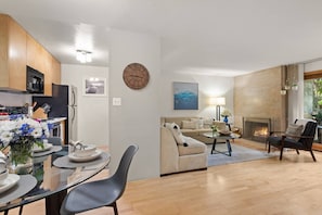 A modern living room with a fireplace, a dining area with a glass table, and a kitchen with wooden cabinets.