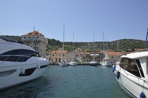 A marina with several yachts docked, a historic building with a red roof, and a hilly landscape in the background.