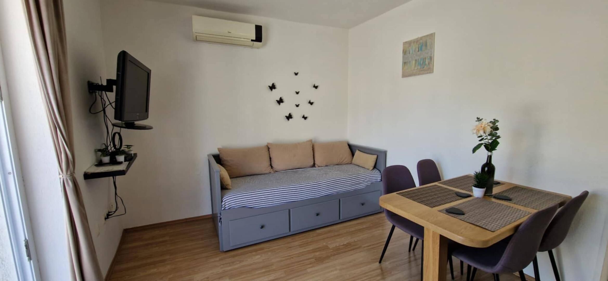 Habitación compacta con sofá cama, televisor montado en la pared, mesa de comedor con sillas y una pequeña planta en maceta.