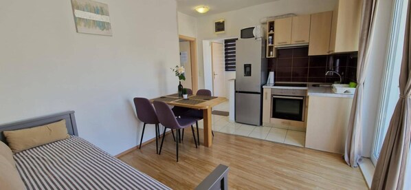 A compact living space with a bed, a dining area with chairs, a small kitchen with a refrigerator and stove, and a window with curtains.