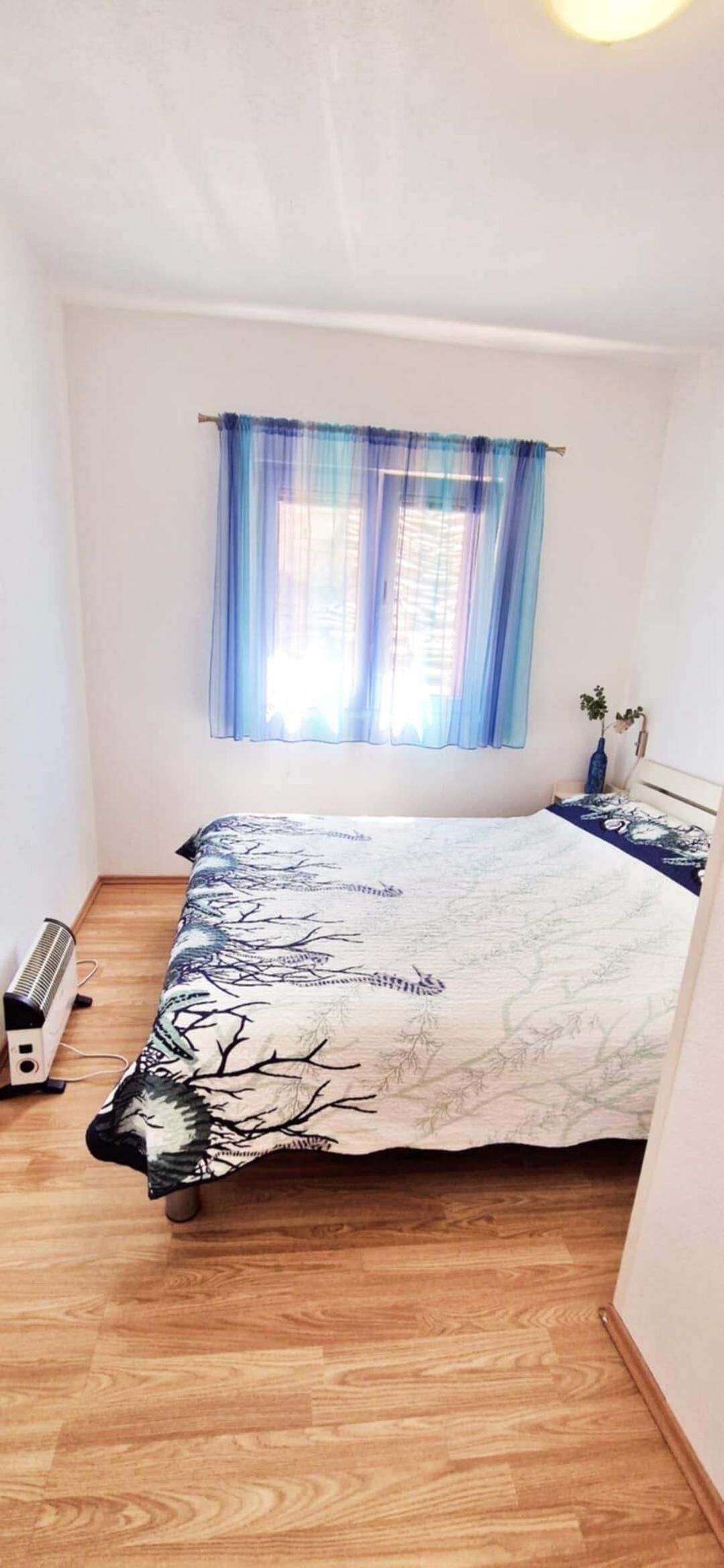 Un dormitorio con cama, cortinas azules en la ventana, una mesita de noche y una planta pequeña.