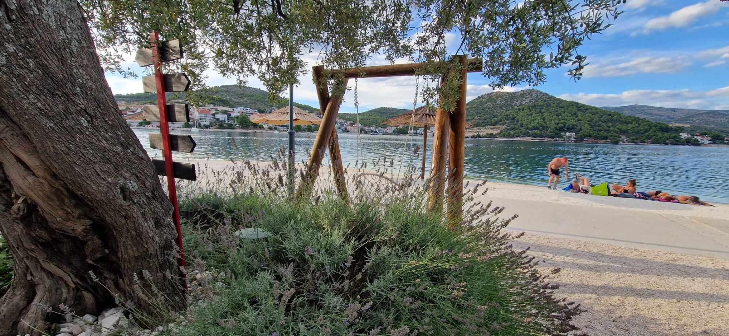 Blick auf einen See mit einer Holzschaukel, einem großen Baum und einem Strandabschnitt, wo Menschen entspannen.