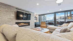 A modern living room with a fireplace, a flat-screen TV, and a large window overlooking a snowy landscape. - Mountain Luxury| 2 King Suites • Fireplace • Private Patio for Après-Ski Bliss (Vail)