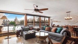 A spacious living room with a large window, a ceiling fan, and a dining area with a chandelier. - Cozy Mountain Home| 3BR• Sleeps 7 • Spacious Mountain Retreat (Vail)