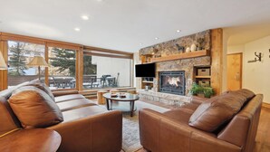 A modern living room with leather sofas, a stone fireplace, and a large window overlooking a deck. - Modern Vail Retreat | King Suite + Bunk Room • Stone Fireplace • Huge Patio (Vail)