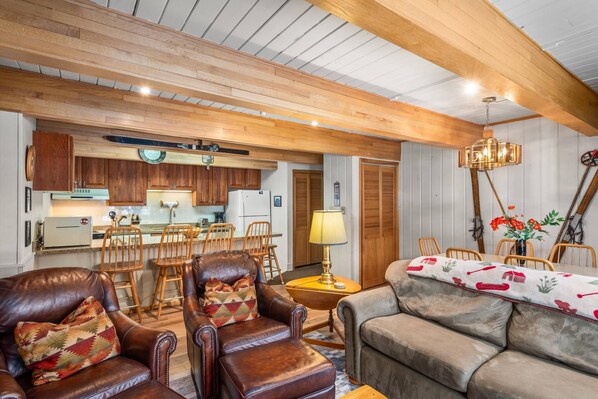 A cozy living room with leather armchairs, a sofa, wooden ceiling beams, a dining area with a table and chairs, and a kitchen in the background.