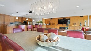 Una cuina moderna amb armaris de fusta, zona de menjador amb taula gran i cadires vermelles, i sala d'estar amb llar de foc i televisió.