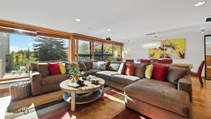 Ein modernes Wohnzimmer mit einer Eckcouch, einem Couchtisch und großen Fenstern mit Blick auf Bäume und Berge.