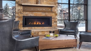 Une cheminée en pierre avec un encadrement noir moderne, deux fauteuils en cuir, une table basse en bois et une vue sur des arbres enneigés à travers de grandes fenêtres.