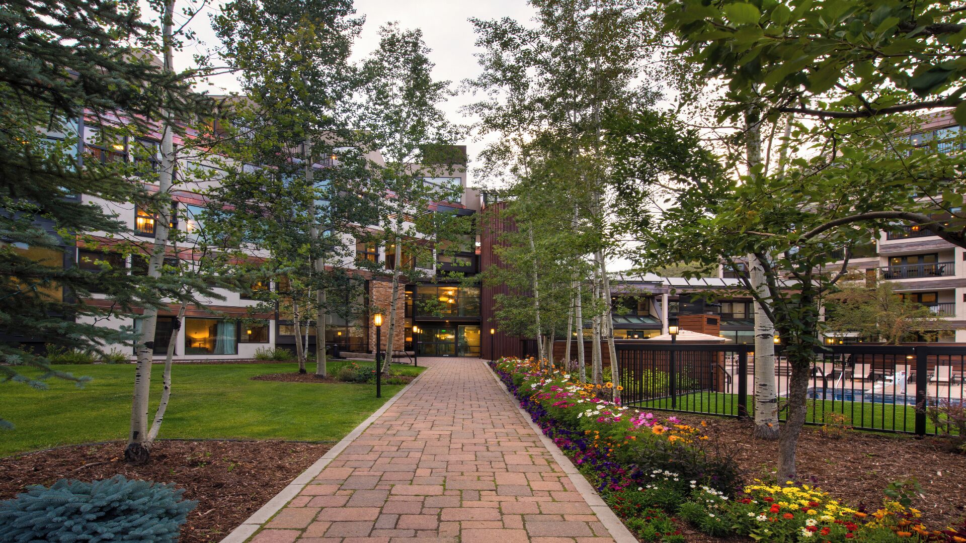 A pathway leading to a building with multiple floors, surrounded by trees and a flower bed.