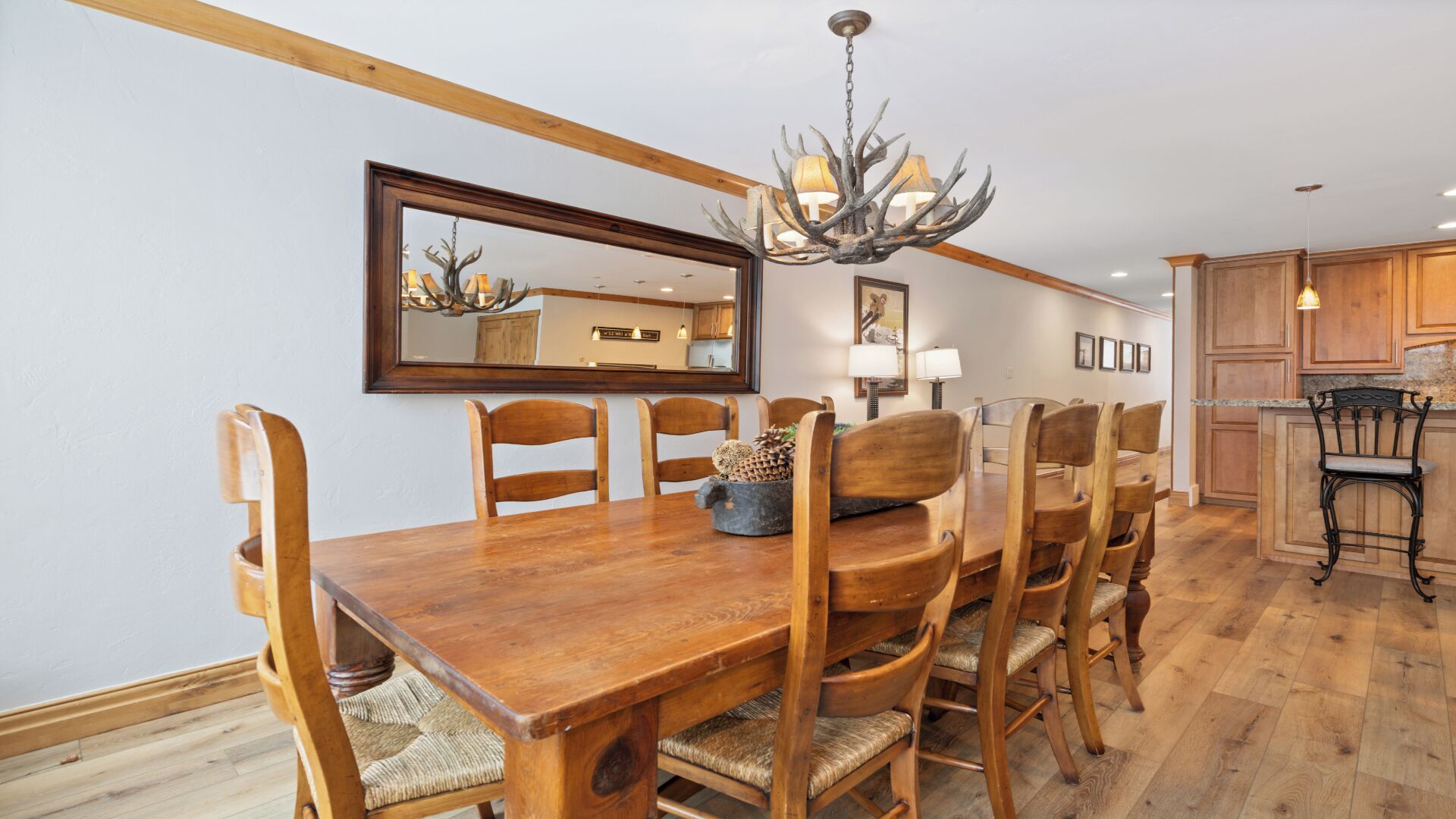 A dining room with a wooden table and chairs, a chandelier, and a kitchen in the background.