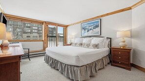 Habitación de hotel con una cama grande, un escritorio, una silla, una mesita de noche, una lámpara, un cuadro en la pared y vistas de edificios a través de la ventana.