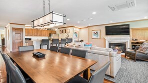 Uma cozinha moderna com armários de madeira, sala de jantar com mesa e cadeiras de madeira, sala de estar com sofá e lareira, e uma TV fixada na parede.