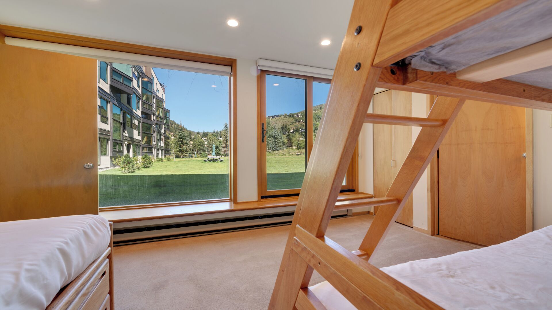 Une chambre avec des lits superposés, de grandes fenêtres et une vue sur un immeuble et la verdure environnante.
