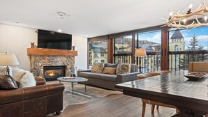 Ein Wohnzimmer mit Kamin aus Stein, einer Couch, einem Couchtisch und einem Essbereich mit Blick auf Gebäude und Bäume.