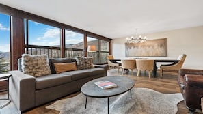 A modern living room with a sofa, coffee table, and dining area, featuring large windows and a mountain view. - Serene 3BR • Stone Fireplace • Gourmet Kitchen & Vail Mountain Views (Vail)