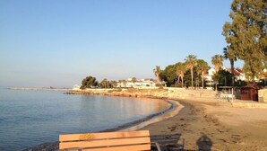 A beach with a wooden bench, calm water, and a row of houses in the background. - ORNIS "Becassina": 1 bedroom+sofa-bed 2/4 pers windows sea view (Sant Carles De La Rapita)