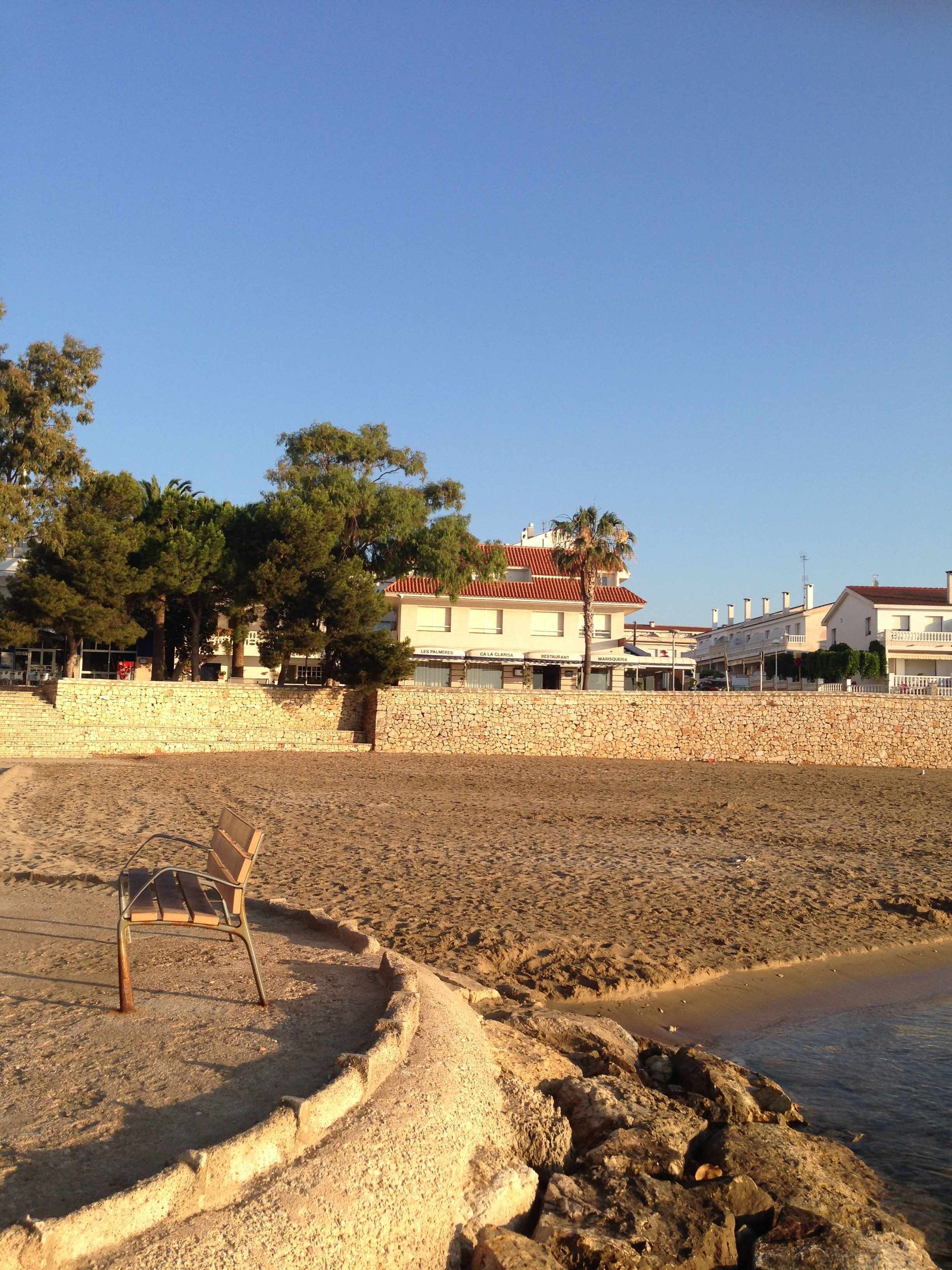 Una playa con un muro de piedra, un banco y edificios al fondo.