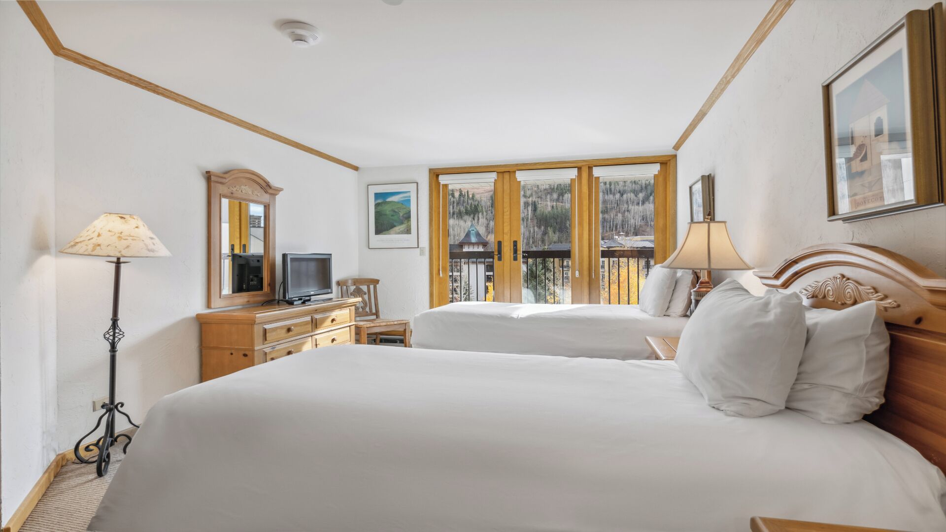 A hotel room with two beds, a desk with a television, and a window with a view of a mountain.