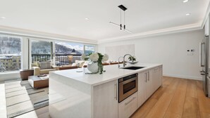 A modern kitchen with a central island, a dining area with a sofa, and a view of snowy mountains through large windows. - Awe-Inspiring 2BR + Den • A/C & Fireplace • Modern Luxury with Stunning Views (Vail)