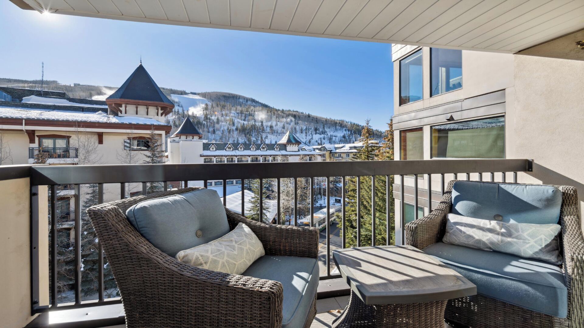 A balcony with wicker furniture, a view of snow-covered mountains, and traditional buildings.
