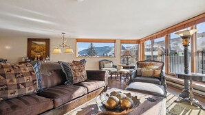 Ein gemütliches Wohnzimmer mit einer Couch, Sesseln, einem Couchtisch und Blick auf Berge durch große Fenster.